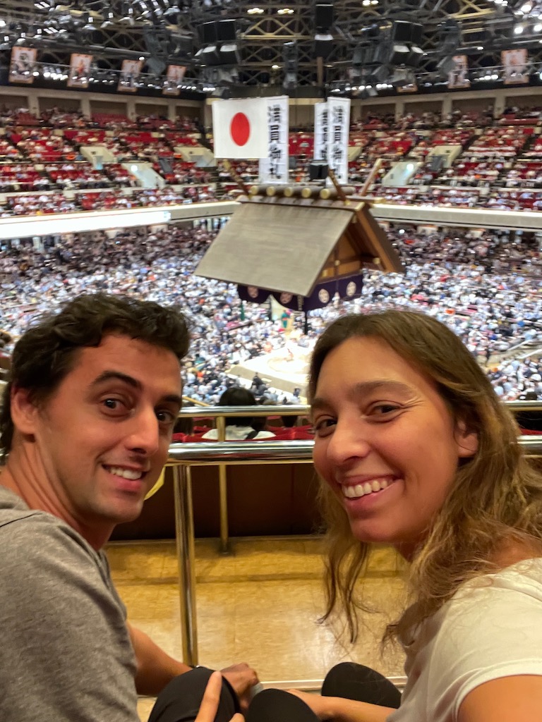 A couple smiling to the camera, with a sumo wrestling stadium behind them