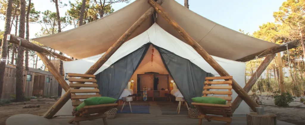 A safari tent surrounded by trees, with two outdoor chairs at the entrance and two beds inside, separated by a wardrobe