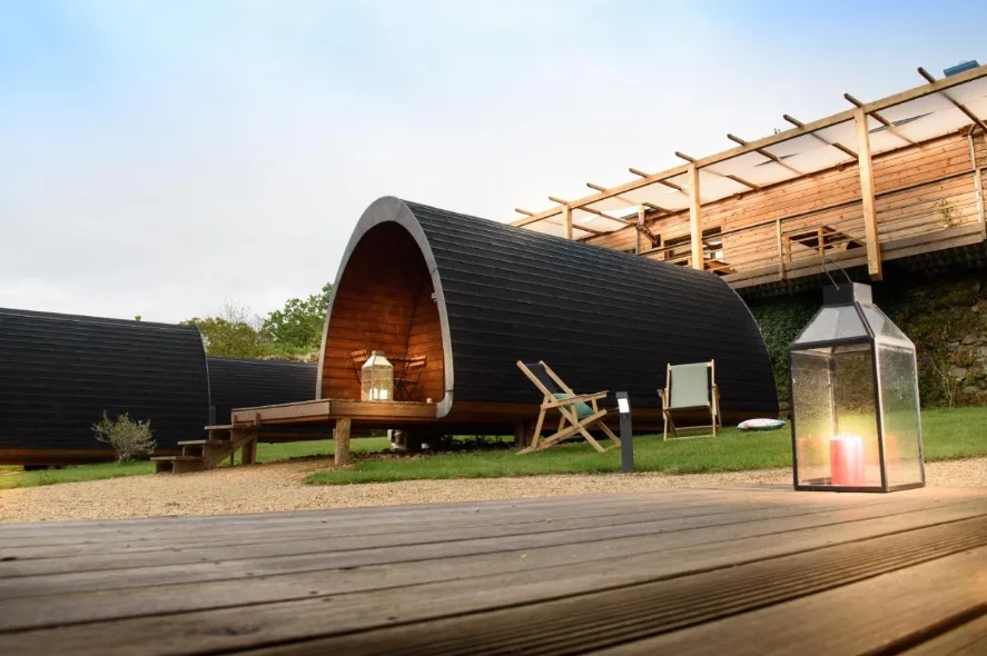 There's a wooden deck on the forefront of the image, with a candle lantern next to it and, in the background, a rounded wooden hut with a porch and outdoor chairs