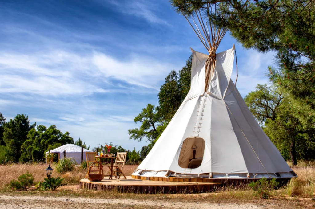 Image of a white tipi tent with two outdoor chairs and a smal table at the entrance, and a glamping tent in the background