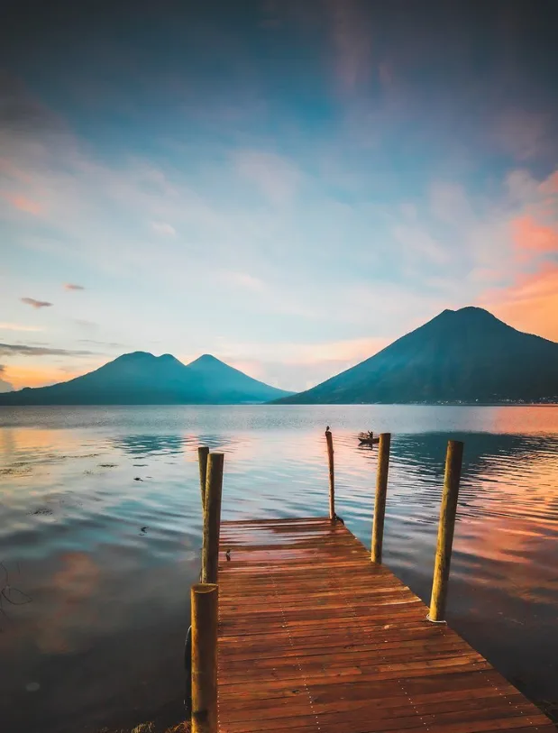 A wooden jetty on a calm lake with two volcanoes in the distance, and pink clouds