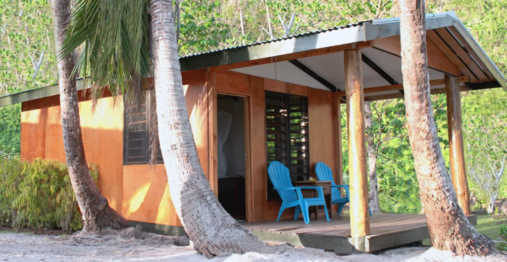 A rustic cabin with a porch and two outdoor chairs, with the trunk of palm trees in front