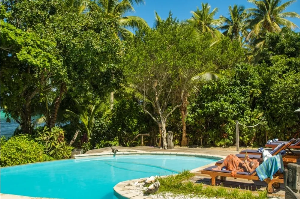 Image of a swimmig pool with lush vegetation in the background, and sunbeds ot the right of the image, where a woman is relaxing with a book