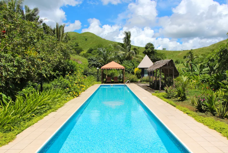 Image of a large swimming pool and a cabana on one end of it, surrounded by vibrantly green vegetation