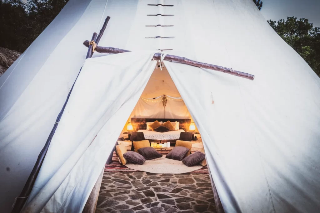 Image of a white tipi tent, and through the open entrance a double bed and cushions on the floor can be seen 