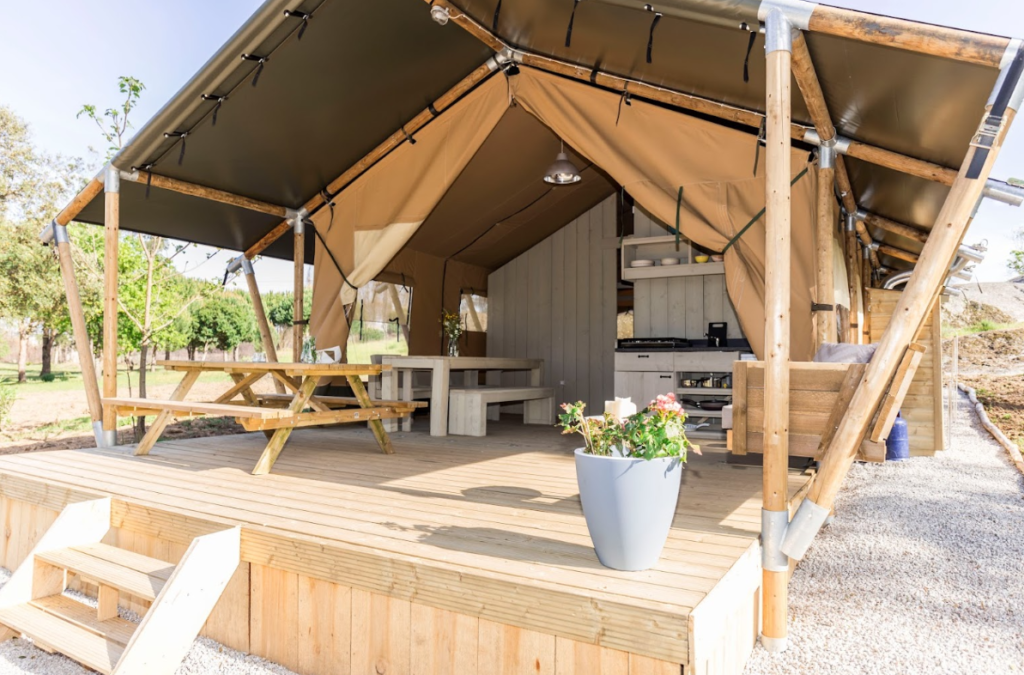 A safari tent with a spacious deck, equipped with outdoor dining table, a flower pot, and a kitchenette.