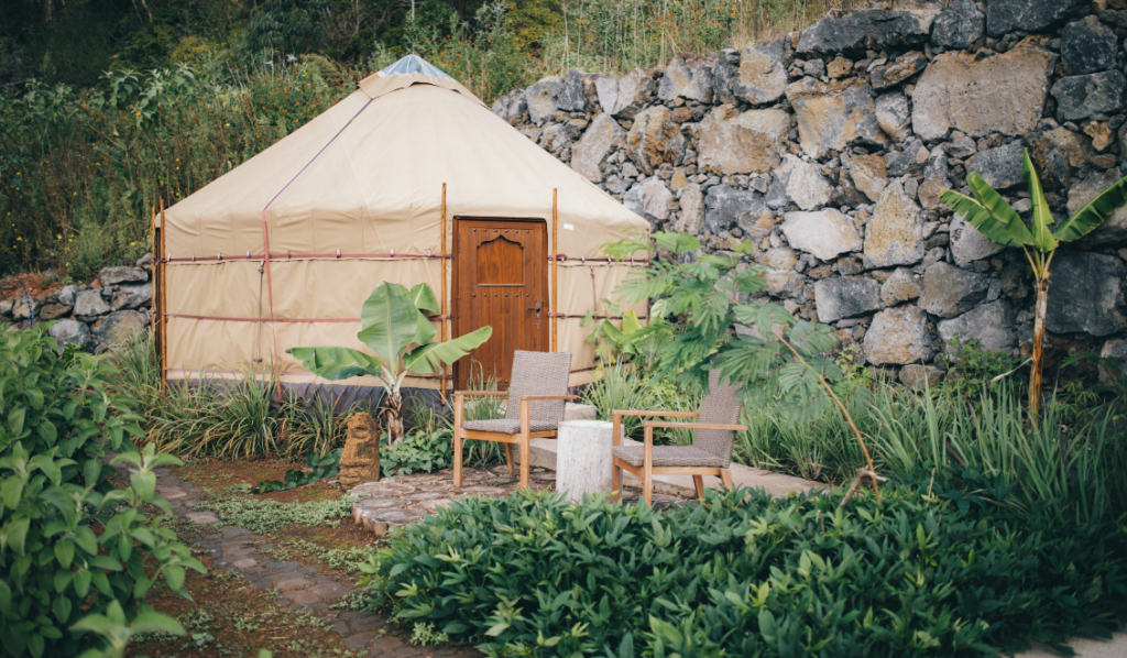 Image of a rounded glamping tent with a wooden door and two chairs at the entrance. To the right there's a stone wall and vegetation all around