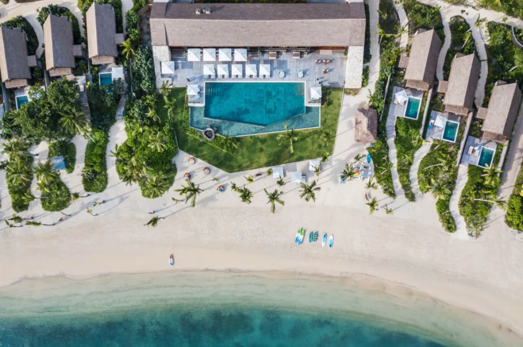 Image of a seaside resort seen from above, with a white sand beach, a large swimming pool and several villas surrounded by palm trees