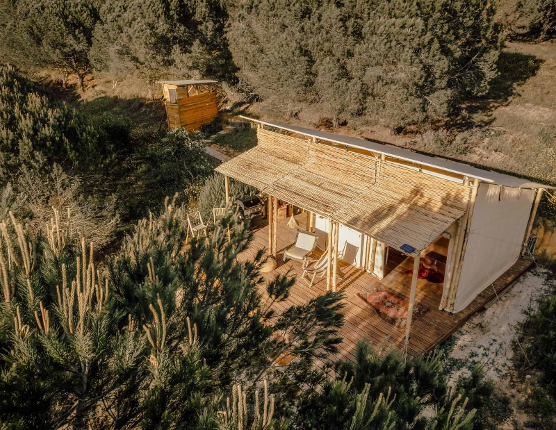 Image seen from above of a safari tent with a wooden porch, and a small shack where the toilet is located to the left of the image, both surrounded by vegetation