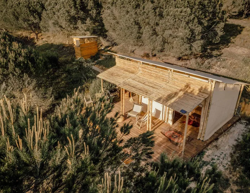 Image seen from above of a safari tent with a wooden porch, and a small shack where the toilet is located to the left of the image, both surrounded by vegetation