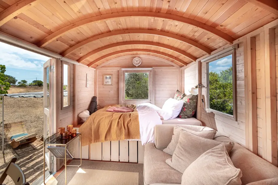 The interior of a cozy hut featuring a double bed surrounded by three windows, a sofa, and an open door leading to the countryside