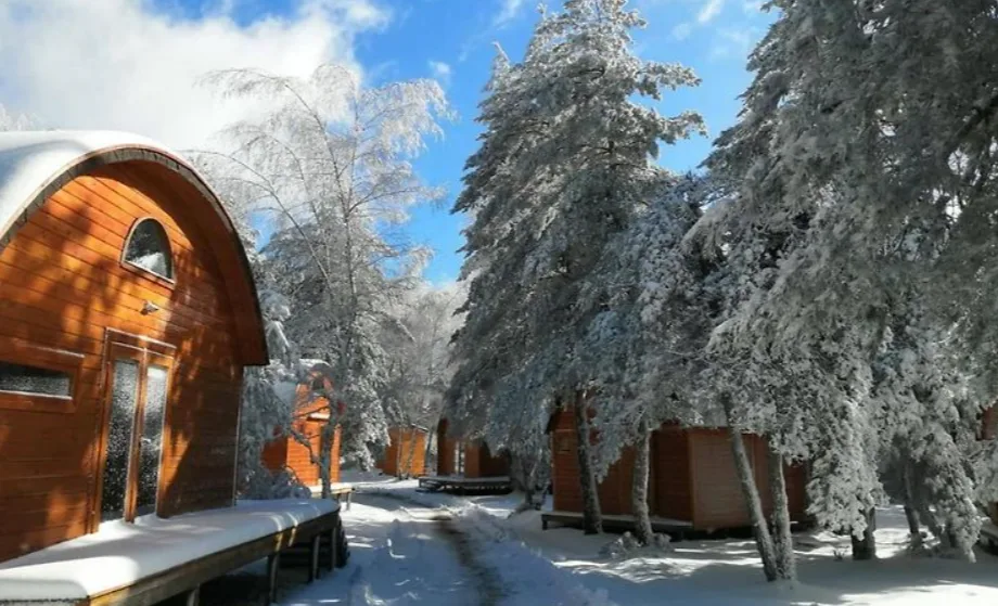 There's a wooden cabin on the left of the image, with its roof and porch covered i snow, and snow-capped trees on the right of the picture