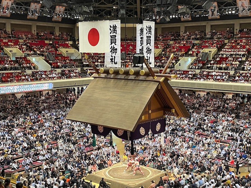 The main stage of a Sumo Wrestling arena, with two wrestlers fighting surrounded by the audience