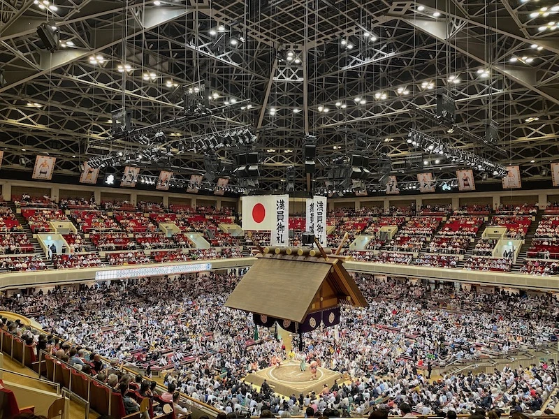 An arena filled with people, and a sumo tournament happening in the main ring