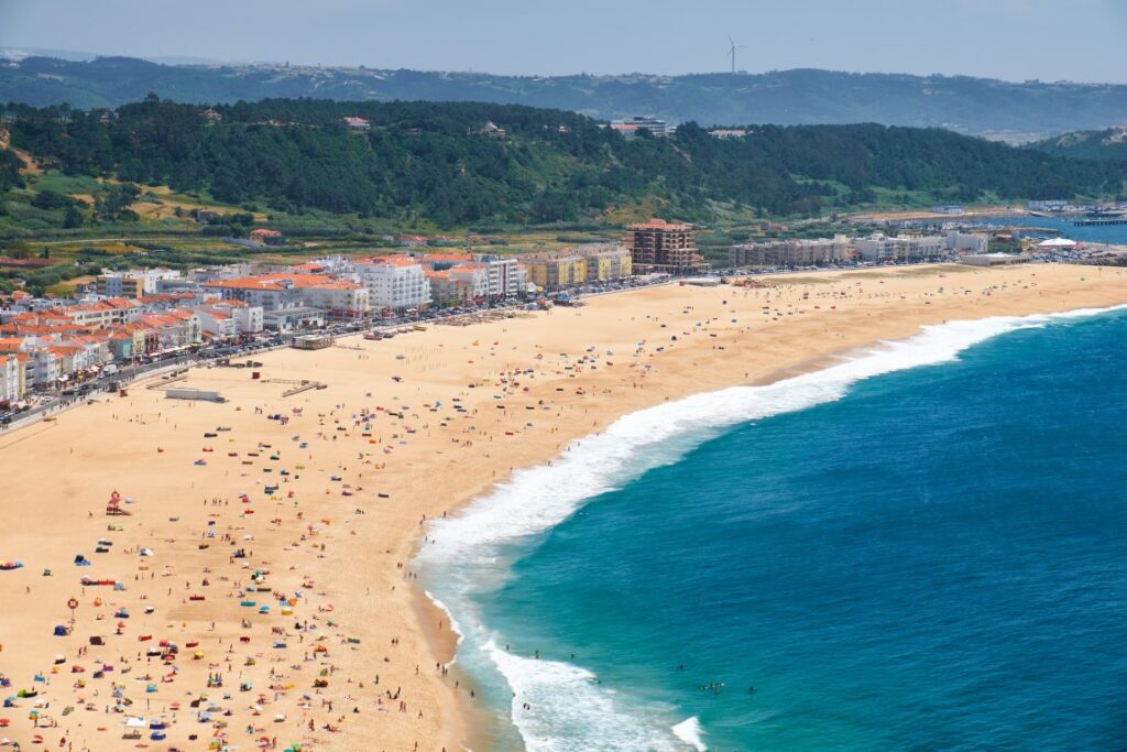 Nazare Beach seen from above, with the Atlantic ocean on the right, the golden coastline dotted with people, and Nazare town on the left