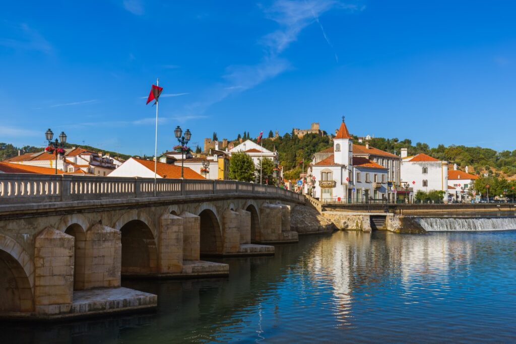 An image of Tomar taken from across the river, with a stone bridge on the left of the picture and white buildings with terracotta-colored roofs on the other side