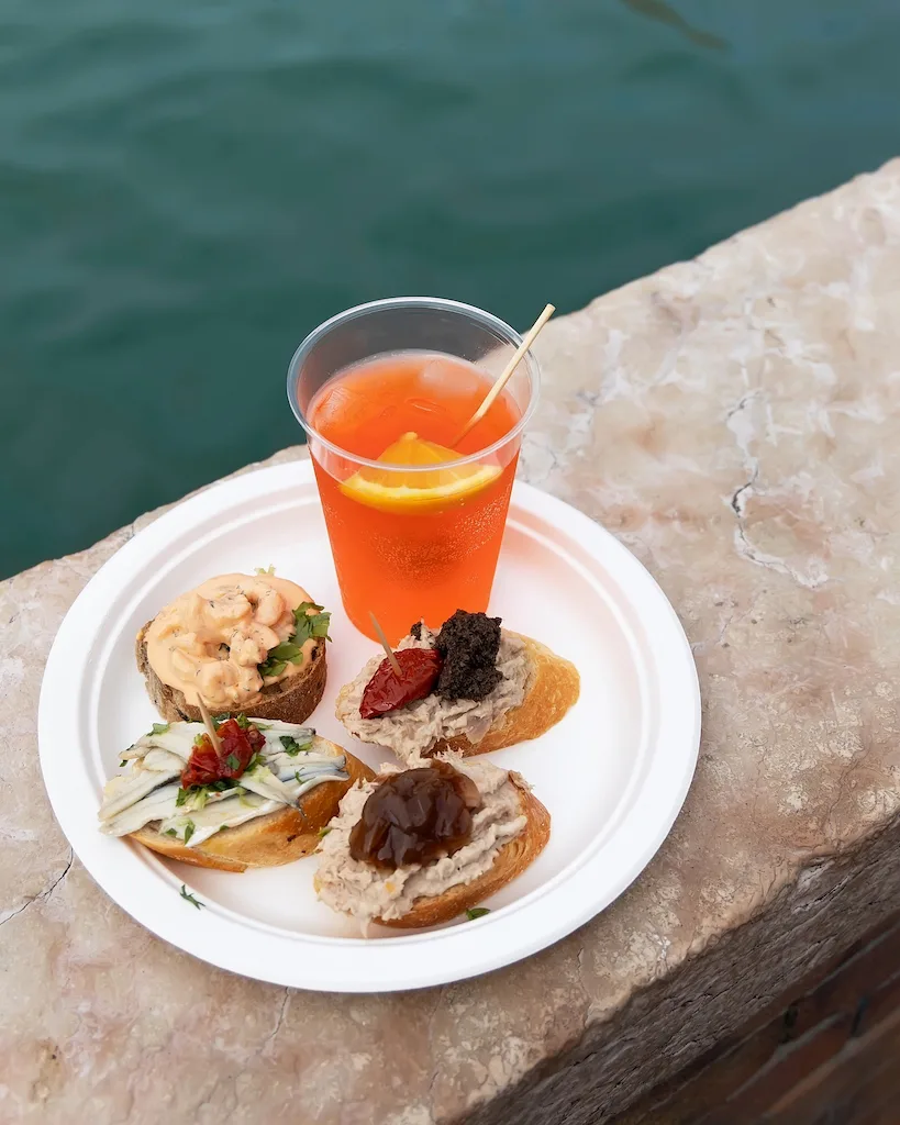 A white paper plate with four bruschetta and an aperol spritz, placed in front of a canal in Venice