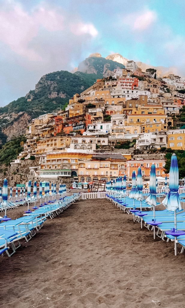 A beach in the Amalfi Coast, with blue sun lounger sand umbrellas on the sandy shore, and a vertical town of colorful houses in the background
