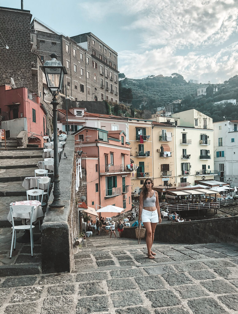A girl walking on the streets of Sorrento, with a staircase on the left, and pastel-colored houses in the background