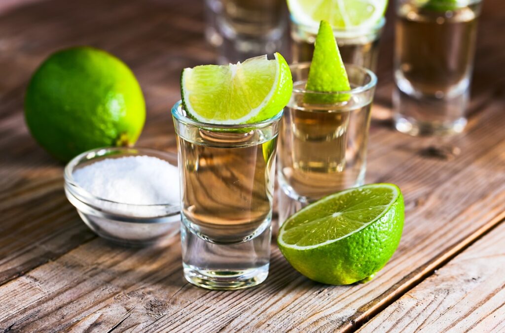 Glasses of tequila, a small bowl of salt, and green limes on a wooden table.