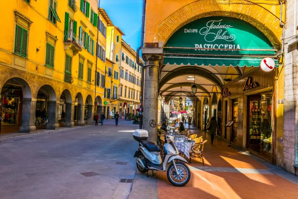 The picturesque Borgo Stretto in Pisa, with its yellow buildings and archways illuminated by the sunlight. To the right of the image there's a covered portico and a grey motorbike parked in front