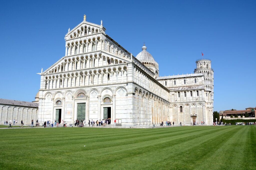 Image of the Cathedral of Pisa taken on a sunny day, with the clear blue sky in the background and green grass surrounding the building