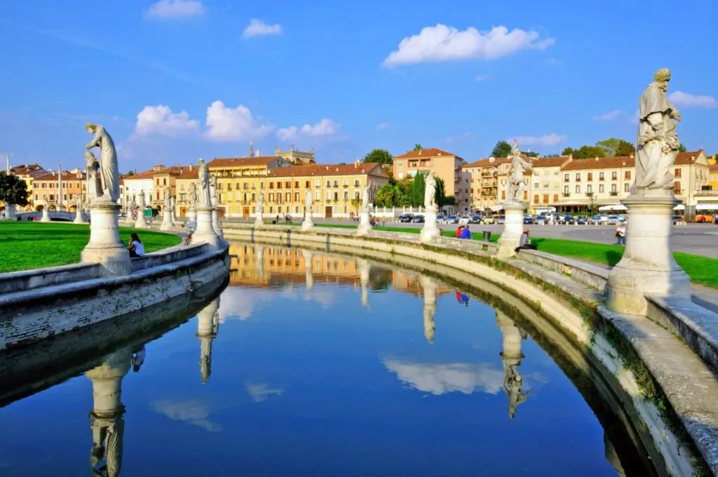 Image of Prato della Valle Square in Padua, featuring one of its waterways lined by statues, and yellow houses with terracotta rooftops in the background