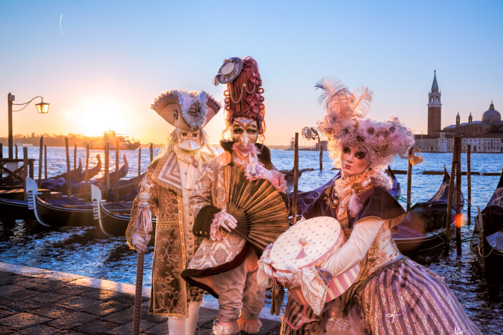 Two men and a woman dressed in elaborate carnival costumes in Venice, with gondolas and a waterway in the background