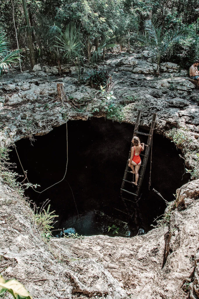 Woman with a red swimsuit standing on a wooden staircase that leads down to a semi-open cenote.