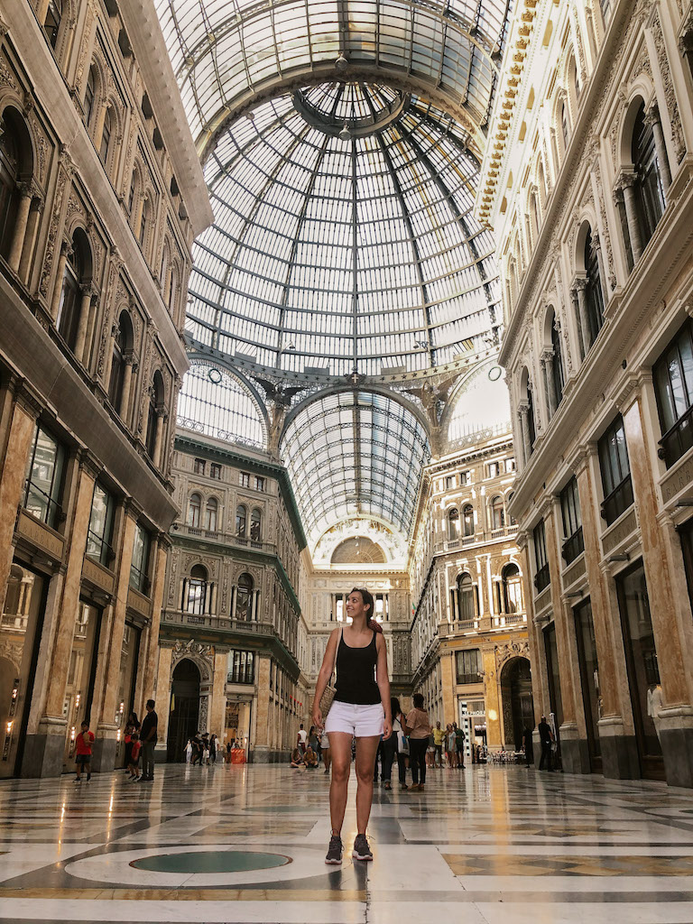 The elegant interiors of Galeria Umberto I in Naples. A woman in a balck tank and white shorts is posing in the center 