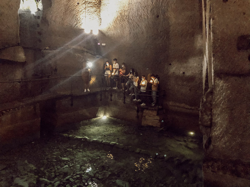 Image of the underground tunnels of Naples, with tour participants standing on the stairs and illuminating the site wirh flashlights