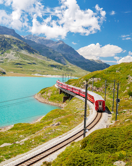 Red bernina train passing through green mountains and by a lake.