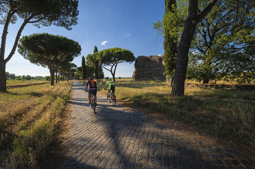 Two people riding bikes along a cobblestone street lined by grass and trees