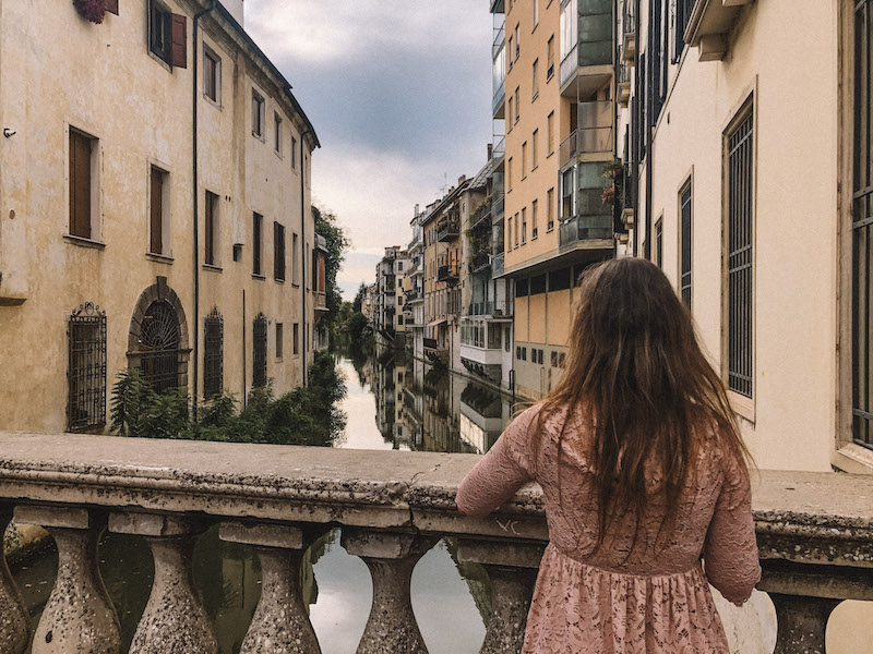 A woman in a pink dress watching a waterway lined by old buildings, inserted in a post about things to do in Padua