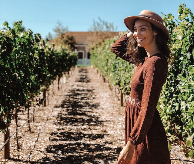 Girl with a brown dress and a brown hat smiling with a vineyard in the backgorund during a wine tour in Verona.