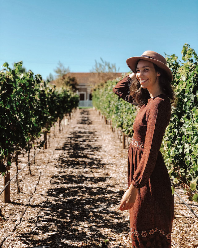 Girl with a brown dress and a brown hat smiling with a vineyard in the backgorund 