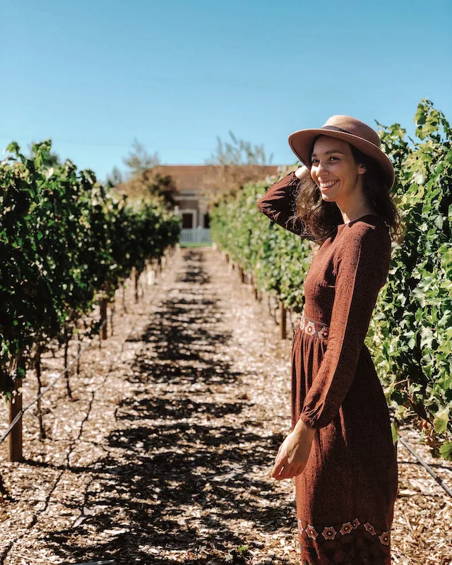 Girl with a brown dress and a brown hat smiling with a vineyard in the background during a wine tour in Verona.