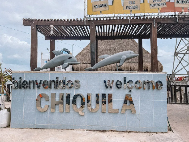 Grey welcome sign to Chiquila Port with two grey dolphin statues on top.