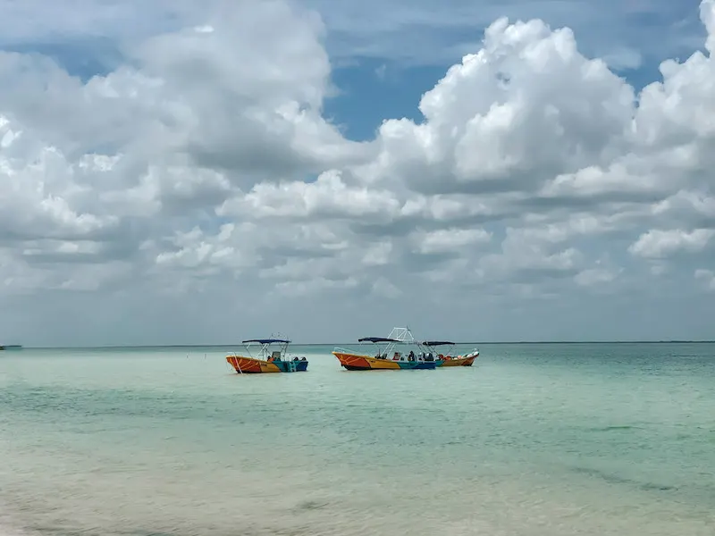 Two red, yellow, green and blue small boats in a turquoise sea under a blue sky, isnerted in a post about how to get from Playa del Carmen to Holbox Island.