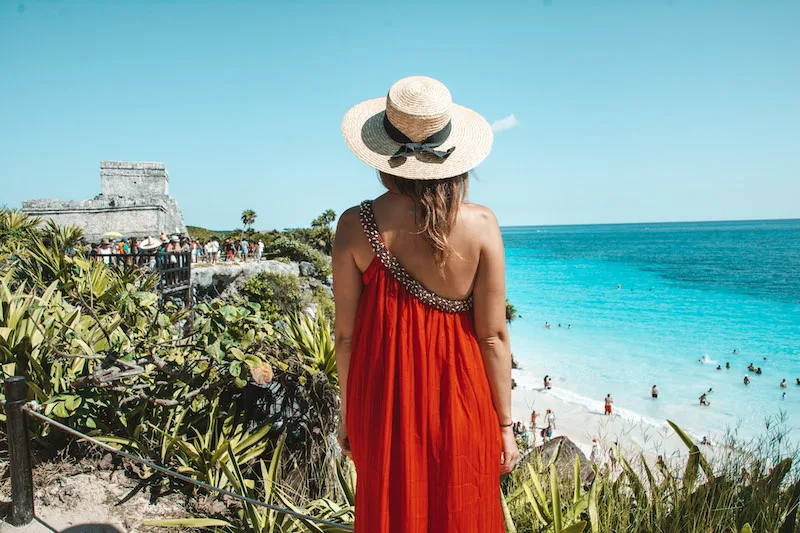 Woman from the back with a red dress and a straw hat standing in front of the Tulum ruins and the sea.