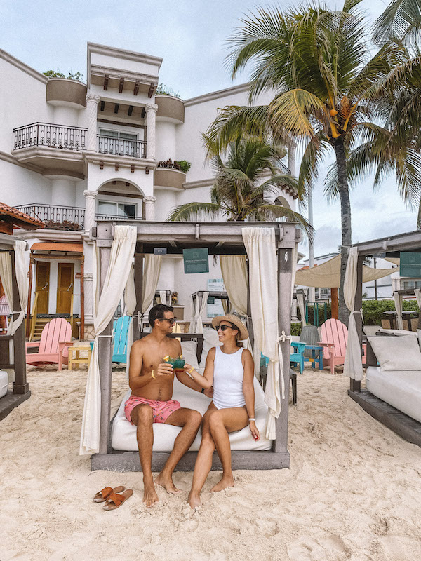 A man and a woman looking at each other and clinking glasse while sitting in a private cabana on the beach in the Riviera Maya.