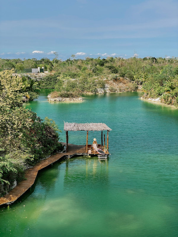 Image of a green-colored lagoon surrounded by lush vegetation, and a woman standing by it