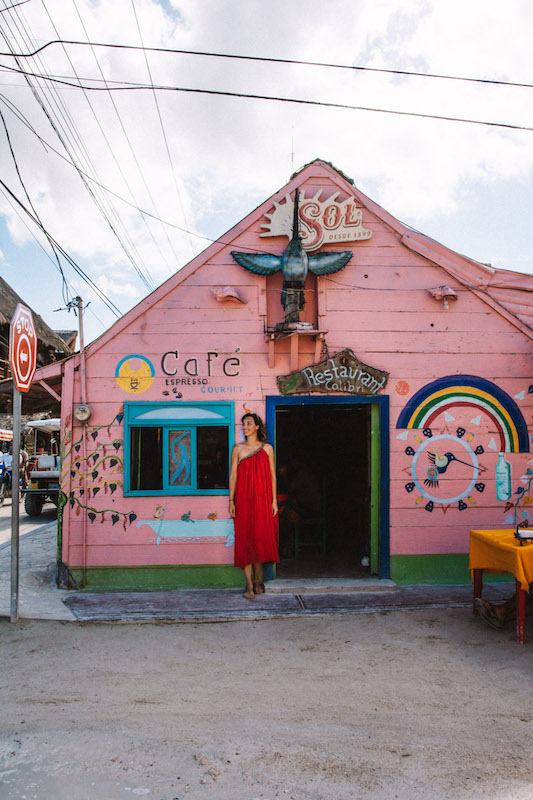 Woman wearing a red dress standing in front of a pink house with murals on the wall.