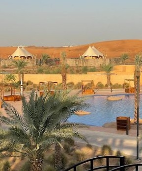 Image of Al Bayader Retreat in Dubai Desert, with a swimming pool surrounded by cabanas and palm trees, and the sand dunes in the bacjground