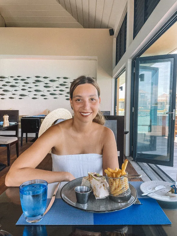 A woman sitting at a table with a plate of fries and a wrap in front of her, smiling at the camera