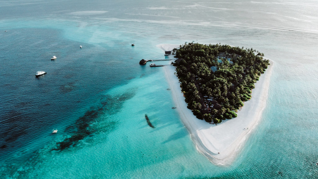 A drone image of a small white-sand island covered in greenery and surrounded by turquoise waters