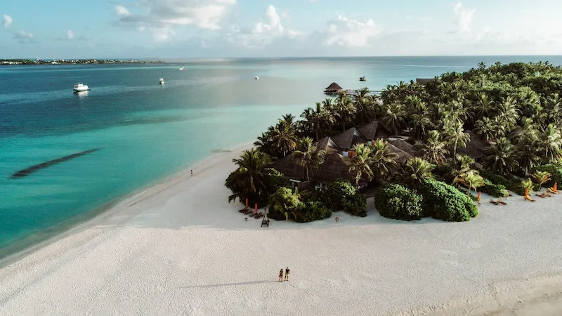 A drone image of a couple standing in a vast beach area with white sands, and the blue sea and lush vegetation in the background