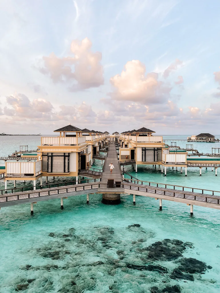 A woman walking on a long wooden jetty over turquoise waters lined by multiple villas on both sides