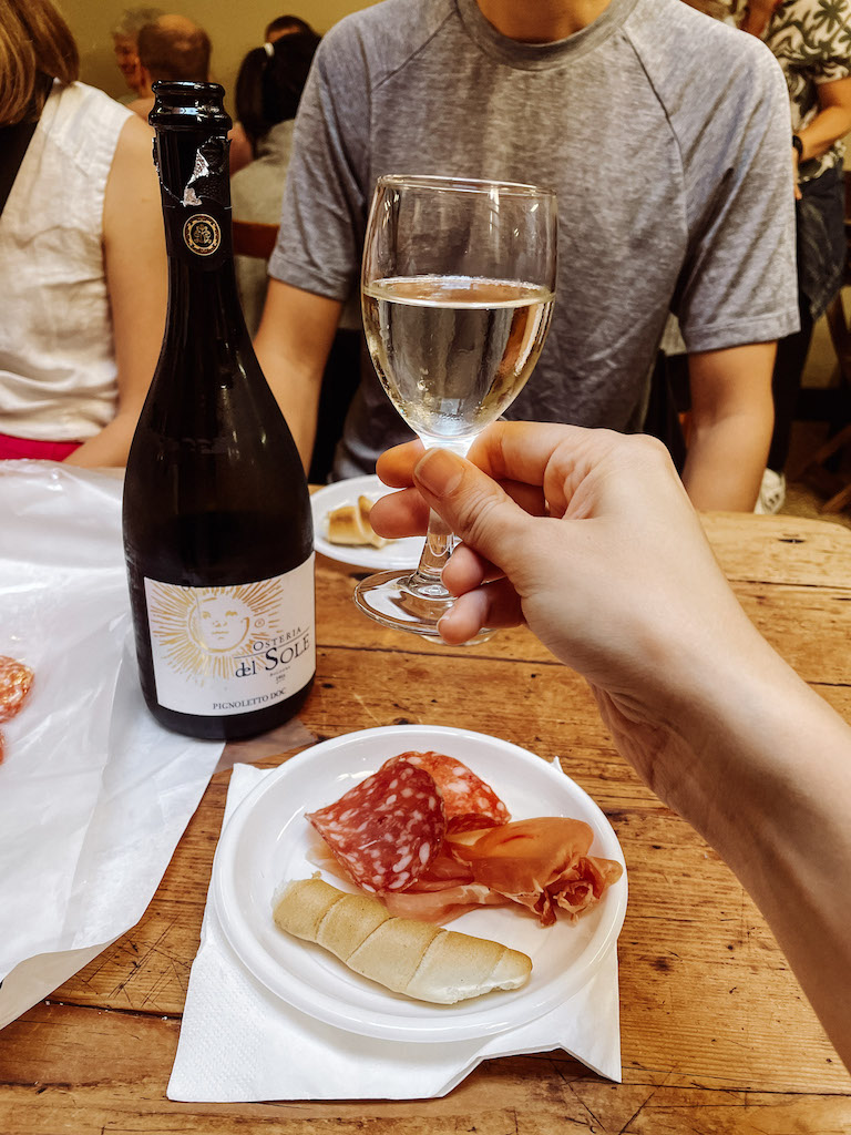 A glass of white wine being held by a woman's hand, and a paper plate with snacks