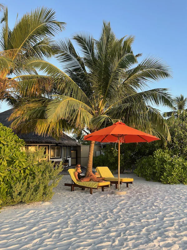 A woman lying on a sunbed in the beach, with an orange sun umbrella next to her and palm trees behind her